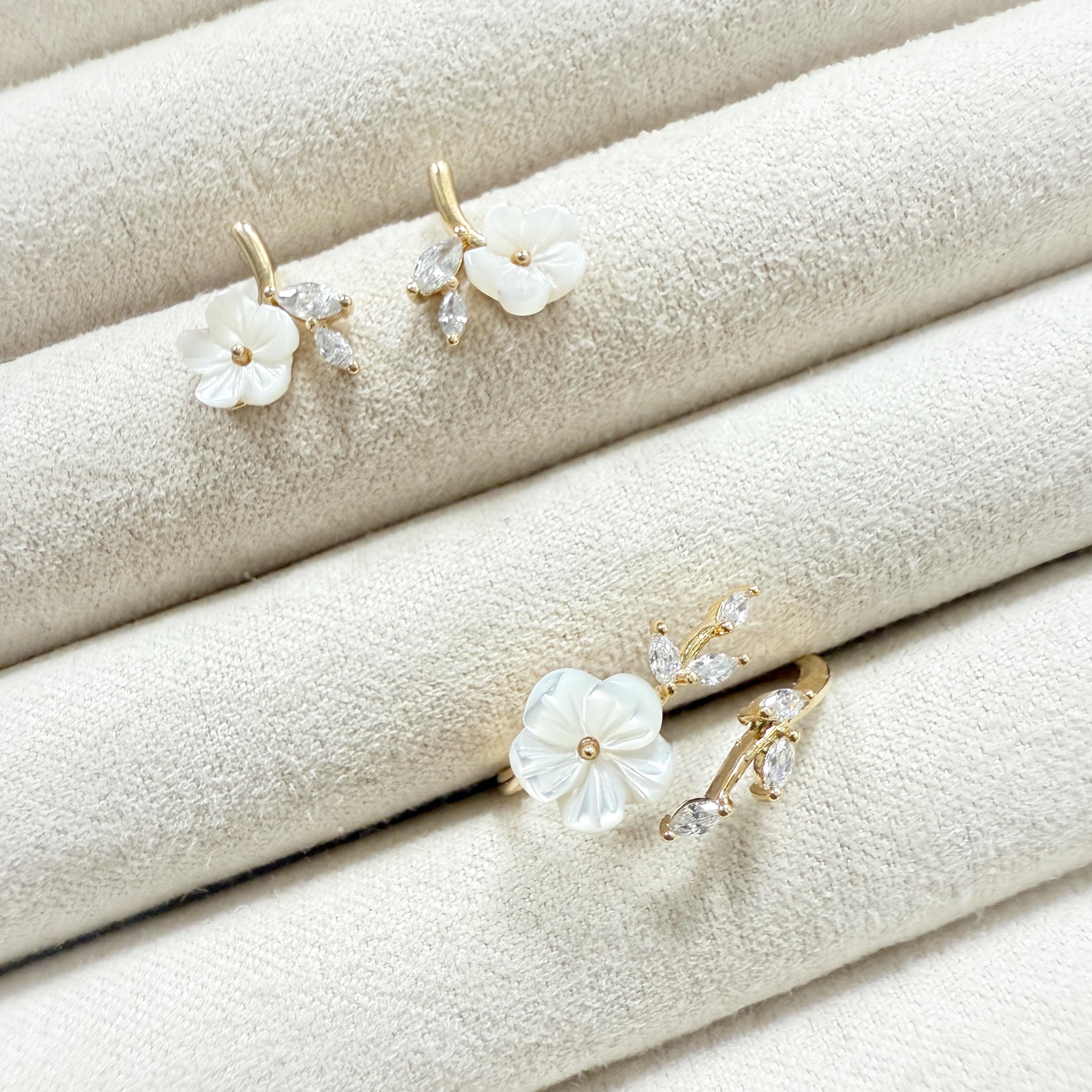 May include: A set of delicate floral jewelry, including a pair of earrings and a ring. The earrings and ring feature white flower-shaped elements with gold-colored stems and accents. Clear, sparkling stones are set on the stems.