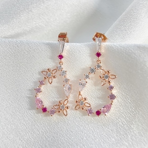 May include: Rose gold dangle earrings featuring a floral design. The earrings are embellished with pink and clear cubic zirconia stones.
