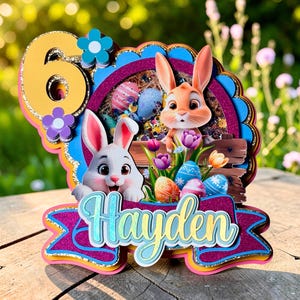 May include: A colorful Easter-themed cake topper featuring two cartoon bunnies, Easter eggs, and the name "Hayden". The number "6" is prominently displayed. The design includes flowers and a wooden fence.