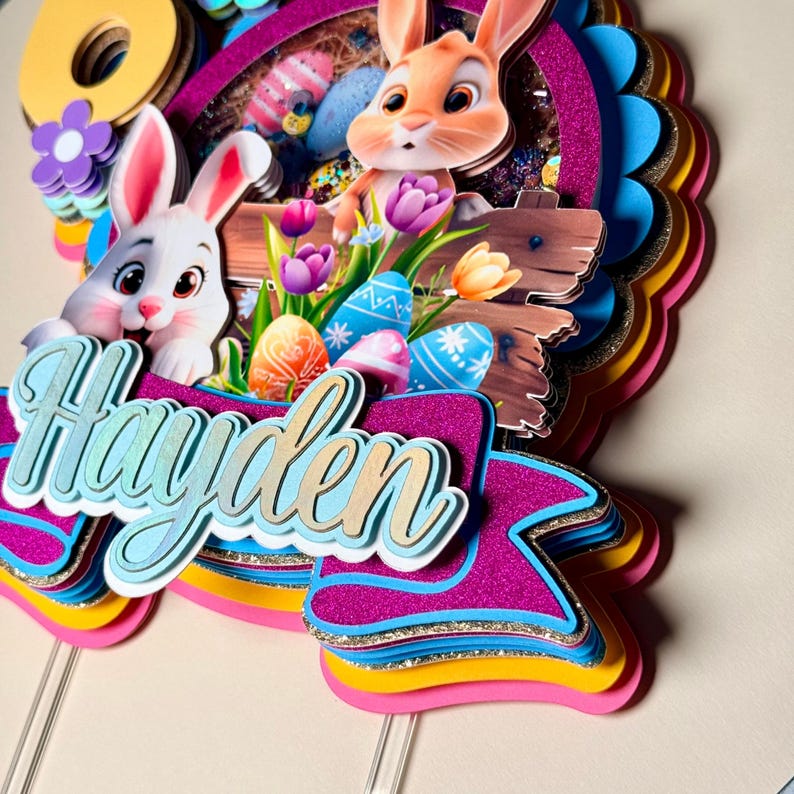 May include: A layered Easter-themed cake topper with the name "Hayden" in a script font. The topper features two cartoon bunnies, Easter eggs, flowers, and a wooden fence. The color scheme includes pink, blue, yellow, and gold glitter accents.