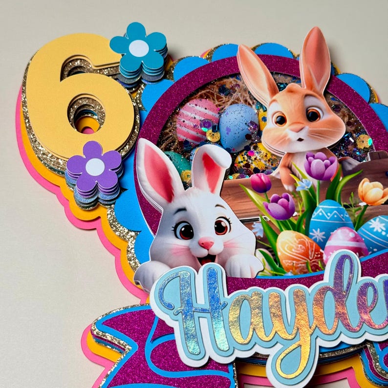 May include: A layered Easter-themed cake topper with the number 6 in yellow and gold glitter. Features two cartoon bunnies, Easter eggs, flowers, and the name "Hayden" in a cursive font. The design incorporates shades of pink, blue, and purple.