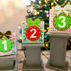 May include: Three decorative Christmas countdown blocks. Each block features a number from 1 to 3 inside a red or green ornament shape, adorned with a bow. The blocks are set on small, gray wooden stands, with a Christmas tree in the background.