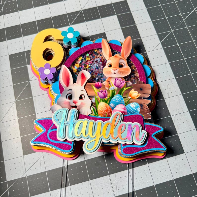 May include: A colorful Easter-themed cake topper featuring two cartoon bunnies, Easter eggs, and the name "Hayden". The number "6" is prominently displayed, along with floral accents. The topper has multiple layers with glitter and vibrant colors.
