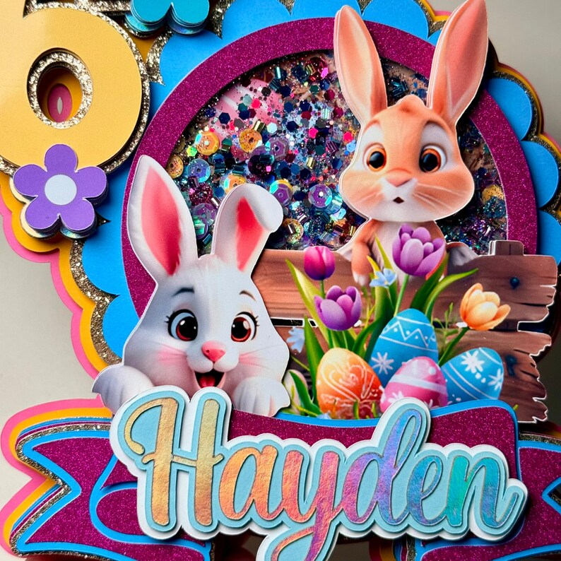 May include: A colorful Easter-themed cake topper featuring two cartoon bunnies, Easter eggs, and flowers. The topper includes the name "Hayden" in a decorative font. The background has a glittery effect with a mix of colors.