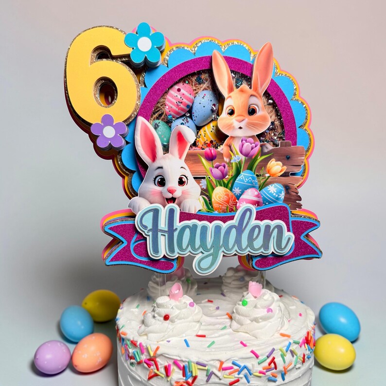 May include: A festive Easter cake topper featuring two cartoon bunnies, colorful Easter eggs, and the name "Hayden" on a banner. The number "6" is also included. The cake is decorated with white frosting and sprinkles, with colorful Easter eggs around the base.