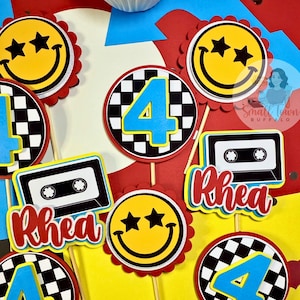 May include: A collection of colorful birthday party decorations. Includes smiley face and cassette tape designs, along with the number 4. The name "Rhea" is also featured. The decorations are on wooden sticks.