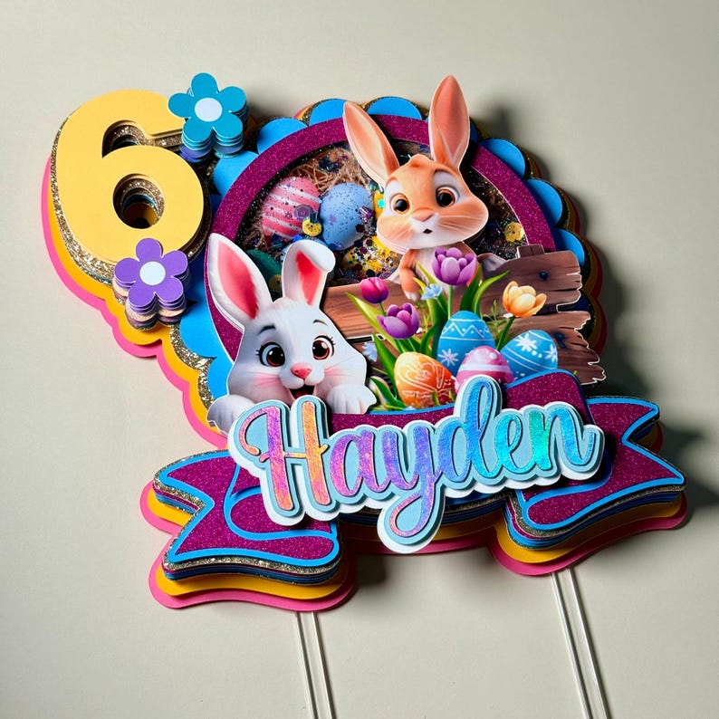 May include: A colorful Easter-themed cake topper featuring two cartoon bunnies, Easter eggs, and flowers. The topper includes the number "6" and the name "Hayden" in a decorative font. The design incorporates shades of pink, blue, yellow, and gold.