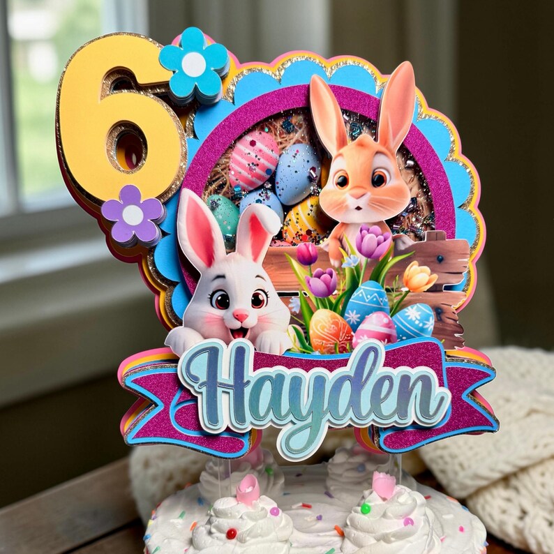 May include: A colorful Easter-themed cake topper featuring two cartoon bunnies, Easter eggs, and the name "Hayden" on a banner. The number "6" is displayed in yellow with floral accents. The topper is designed for a birthday cake.