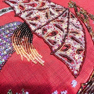 May include: Close-up of a red fabric with a beaded design. The design features a large, intricate pattern of leaves and flowers, with iridescent beads in shades of purple, pink, and gold.