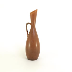 May include: A brown ceramic vase with a handle. The vase has a slender, elongated body that widens towards the base. The top is angled, and the handle is a curved loop. The vase is a warm brown color with a textured surface.