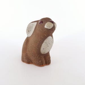 Könnte beinhalten: Eine kleine Keramikfigur eines Kaninchens mit brauner und weißer, gesprenkelter Glasur. Das Kaninchen hat rote Augen und sitzt aufrecht mit hochgestellten Ohren.