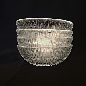 May include: A stack of four clear glass bowls with a textured, vertical-line pattern. The bowls are stacked on top of each other, with the top bowl slightly overlapping the one below. The background is a solid black.