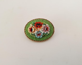 May include: Oval mosaic brooch featuring a floral design. The brooch has a green background with a gold-colored border. The flowers are red, white, blue, and pink. A vintage-style accessory.