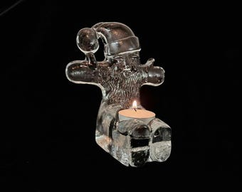 May include: A clear glass Santa Claus figurine with a lit tea light candle. The Santa figure has a hat, beard, and outstretched arms. The candle sits in a holder at the base of the figurine. The background is black.