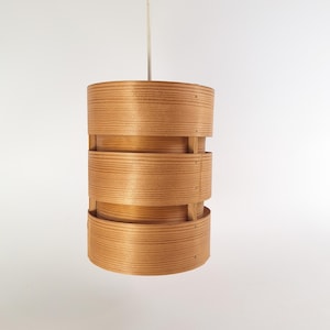 May include: A wooden pendant light with a cylindrical shade made of stacked, thin wood slats. The shade has a natural wood finish and is suspended from a white cord.