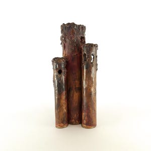 May include: A set of three copper vases with a textured, rustic finish. The vases are arranged in a staggered formation, with the tallest vase in the center. Each vase has a unique, irregular shape and a slightly different color.