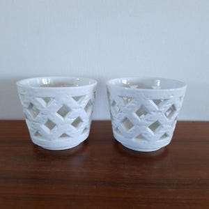 Puede incluir: Dos portavelas de cerámica blanca con un diseño de celosía de diamantes. Los portavelas son cilíndricos con una parte superior ligeramente ensanchada y se asientan sobre una superficie de madera oscura. El fondo es una pared blanca lisa.
