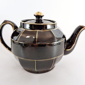 Op de afbeelding: Een zwarte theepot met gouden versiering. De theepot heeft een geometrisch ontwerp en een gouden knop op de deksel.