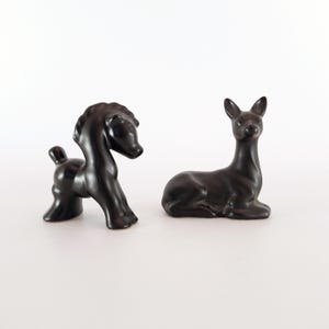May include: Two black ceramic animal figurines, a horse and a deer. The horse is standing on all four legs with its head turned to the side. The deer is lying down with its head raised. Both figurines are made of a smooth, matte black ceramic.