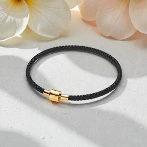 Könnte beinhalten: Schwarz geflochtenes Armband mit goldfarbenem Magnetverschluss. Das kreisförmige Armband liegt auf einer weißen Oberfläche, mit unscharfen Blumen im Hintergrund. Das Design des Armbands ist schlicht und elegant.