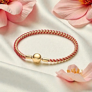 May include: A gold bracelet with a red woven design. The bracelet has a round gold clasp in the center. The background features pink flowers and a white satin fabric.