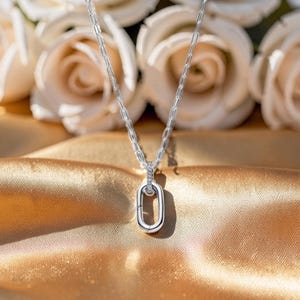 Op de afbeelding: Een zilveren ketting met een rechthoekige hanger, tegen een achtergrond van witte rozen en gouden satijn. De hanger heeft een klein, ovaalvormig ontwerp met een kleine edelsteen. De ketting heeft een delicate schakelstijl.