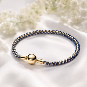 May include: A circular bracelet featuring a braided design of metallic gold and vibrant blue cord. The bracelet is secured with a polished, spherical gold-toned bead clasp that connects the two ends of the braided band into a continuous loop.