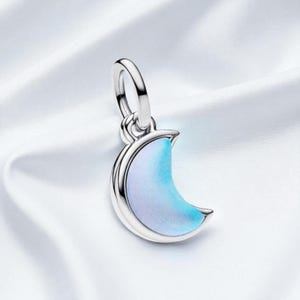 Può includere: Un ciondolo a forma di mezzaluna color argento con finitura smaltata azzurra e bianca. Il ciondolo ha un anello nella parte superiore per il fissaggio a una collana o un braccialetto. Lo sfondo è un tessuto bianco e setoso.