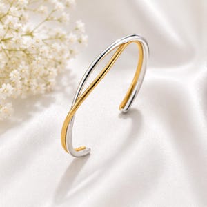 Op de afbeelding: Een tweekleurige armband met een gepolijste zilverkleurige band die buigt en overlapt met een gladde goudkleurige band. Het open ontwerp creëert een strakke, gelaagde uitstraling met een hoogglanzende metallic afwerking over het hele sieraad.