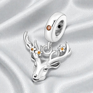 May include: A silver-colored charm featuring a deer head with small orange flower accents on its antlers. The charm has a loop at the top for attaching to a bracelet or necklace. The background is a soft, white, silky fabric.