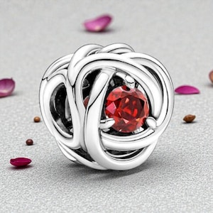 Puede incluir: Un charm de color plateado con un diseño tejido, con una gema roja vibrante en el centro. El charm es redondo y detallado, con un acabado pulido. Alrededor hay pequeños pétalos y motas.
