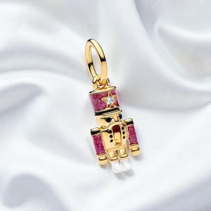 May include: Gold-coloured charm with a miniature nutcracker soldier design. The charm has a red and black hat with a star and a clear gem. The soldier's uniform is gold with red accents and white boots. A festive holiday-themed accessory.
