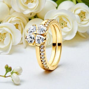 May include: A gold-colored ring set featuring a four-leaf clover design with clear, sparkling stones. The band is adorned with smaller, clear stones. The background includes white flowers.