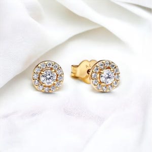 May include: A pair of gold earrings with a round halo setting featuring a single diamond in the center and smaller diamonds surrounding it.