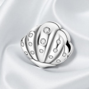 May include: A silver seashell charm with sparkling clear gemstones. The charm is detailed with a shell design and small, round gemstones. The charm is displayed on a white, silky background.