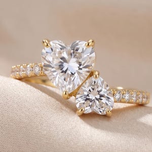 May include: A gold ring featuring two heart-shaped, clear gemstones. The larger heart-shaped stone is positioned above a smaller one. The band is adorned with smaller, round gemstones. The ring is set against a neutral background.