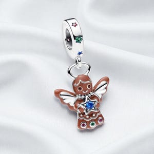 Puede incluir: Un charm de plata con un diseño de ángel de pan de jengibre. El ángel tiene glaseado marrón, alas blancas y una estrella azul. La parte superior del charm tiene detalles de estrellas rojas, verdes y azules. El charm está suspendido de un anillo plateado.