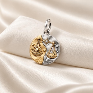 Peut inclure: Un pendentif circulaire bicolore présentant un design divisé avec un soleil doré à gauche et une lune argentée à droite. Le centre représente un motif de balance orné de petites étoiles et d'une pierre claire unique au sommet.