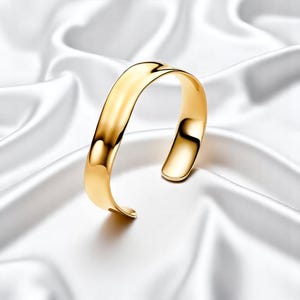 Puede incluir: Un brazalete dorado con un acabado liso y pulido. El diseño abierto se curva con gracia, reflejando la luz. El brazalete se exhibe sobre una tela blanca y sedosa, resaltando su elegante forma. Una pieza de joyería simple pero llamativa.