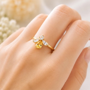 May include: A gold-toned ring featuring a bee-shaped design. The center of the bee is a pear-shaped yellow gemstone, while the wings are accented with two clear marquise-cut stones and a small round clear stone at the head of the bee.