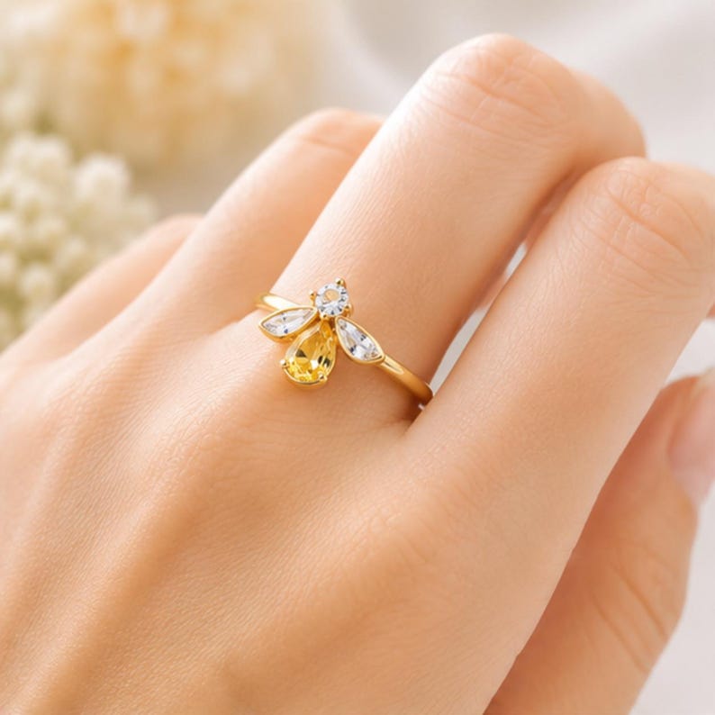 May include: A gold-toned ring featuring a bee-shaped design. The center of the bee is a pear-shaped yellow gemstone, while the wings are accented with two clear marquise-cut stones and a small round clear stone at the head of the bee.