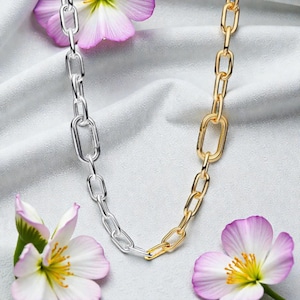 Puede incluir: Un collar de cadena bicolor con eslabones plateados y dorados. El collar se muestra sobre una superficie de satén blanco, con flores rosas y moradas en el fondo. Los eslabones de la cadena tienen forma ovalada y están conectados.