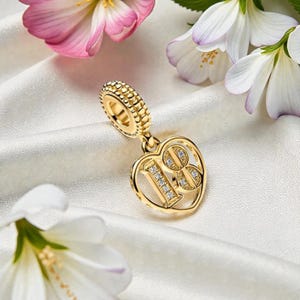 May include: A gold-colored charm with the number "18" in a heart shape, embellished with small, clear stones. The charm hangs from a textured, gold-colored ring. The background features white fabric and flowers.