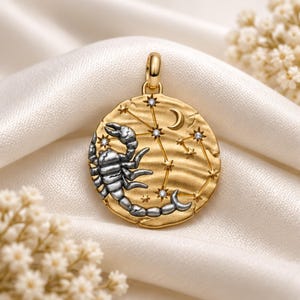 May include: A round gold pendant featuring a raised silver scorpion figure on the left side. The surface displays a textured finish with a celestial design including a crescent moon and a constellation of small stars accented with clear stones.
