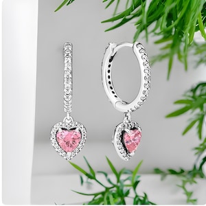 Puede incluir: Un par de pendientes de aro de plata con un colgante de corazón rosa. Cada colgante de corazón está rodeado por un halo de piedras transparentes más pequeñas.