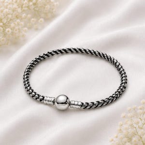May include: A silver-toned bracelet featuring a braided, rope-like chain design. The bracelet includes a central, smooth, spherical bead flanked by two cylindrical, ribbed metal accents that serve as a clasp mechanism for the circular band.