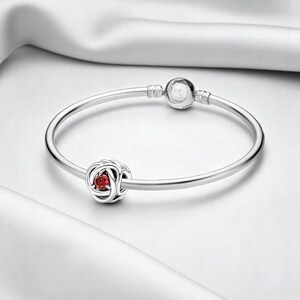 May include: A silver bangle bracelet with a red gemstone charm in the shape of a knotted rose.