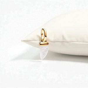May include: A gold-colored pendant with a white, horn-shaped stone. The pendant is hanging from a gold loop. The pendant is resting on a white pillow.