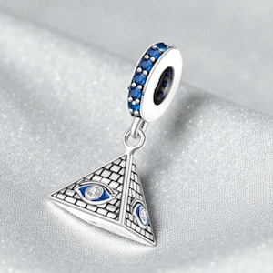 May include: A silver charm with a pyramid design, featuring an eye symbol with blue and white accents. The charm has a ring adorned with blue stones. The pyramid has a brick pattern and is suspended from the ring.