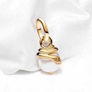 May include: A gold pendant with a white, pearl-like orb. The pendant has a coiled snake design around the orb, with small diamond accents. It hangs from a gold loop, designed for a necklace or bracelet. The pendant is a piece of jewellery.
